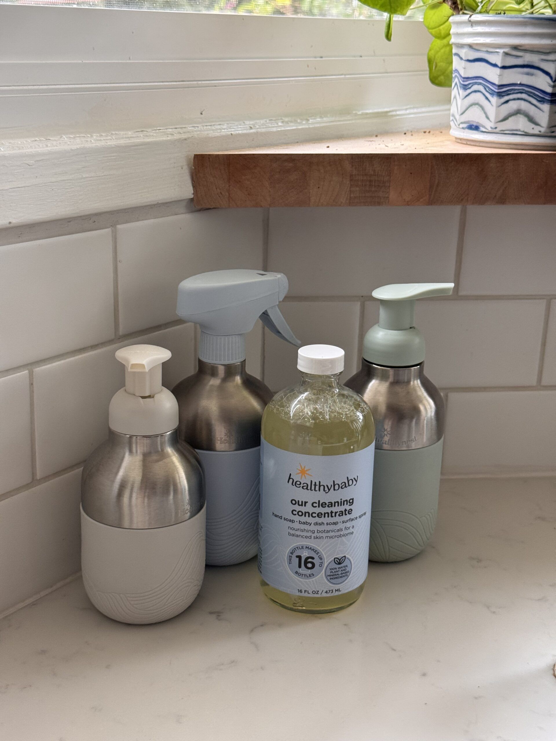 Four cleaning product bottles, including a labeled "healthybaby our cleaning concentrate," are arranged on a white kitchen counter near a tiled wall and a window with a potted plant.
