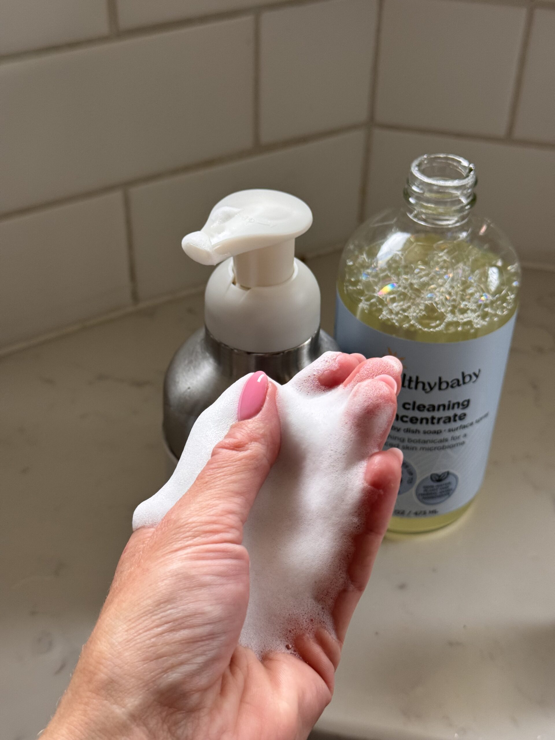 A hand with pink nails holds white foam near a soap dispenser and a bottle labeled “healthy baby Cleaning Concentrate” on a bathroom counter.