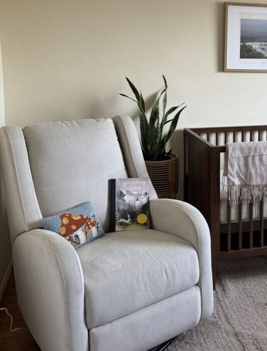 A cream recliner and a wooden crib with a blanket are in a nursery room. A potted plant, small bookshelf, and framed photo are arranged nearby.