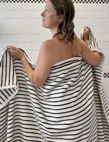 A person with wet hair stands wrapped in a black and white striped organic towel in a tiled bathroom, facing left.