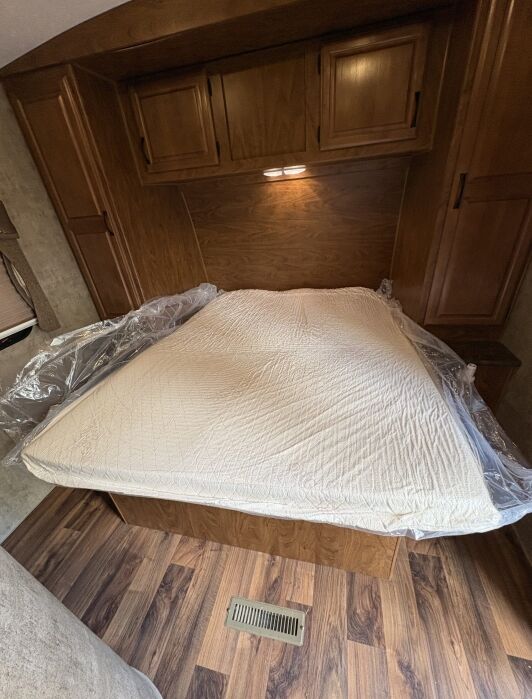 A neatly made bed with a plastic cover partially removed, surrounded by wood cabinets and flooring in a small room.