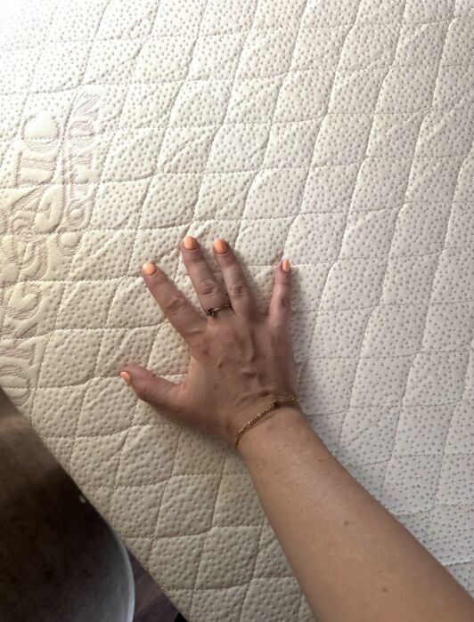 A hand with peach-colored nails presses down on a quilted, light-colored mattress surface with a subtle embossed pattern.