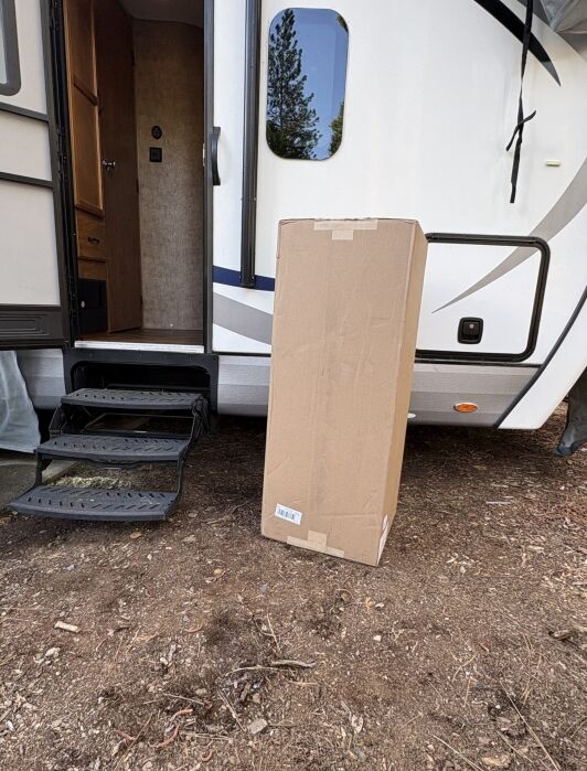 A large cardboard box is standing upright on the ground next to the steps of an RV with its door open.