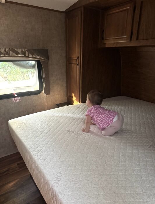 A baby in a pink outfit crawls on a mattress inside an RV, facing a window with a view of greenery outside.