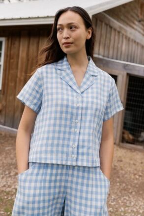 A person stands outdoors in front of a wooden building, wearing a light blue and white checkered short-sleeve button-up shirt and matching shorts, with hands in pockets.