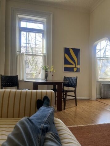 Person lying on a striped sofa with feet up, facing a dining table, two chairs, a vase with flowers, a window, and modern art on the wall in a bright living room.