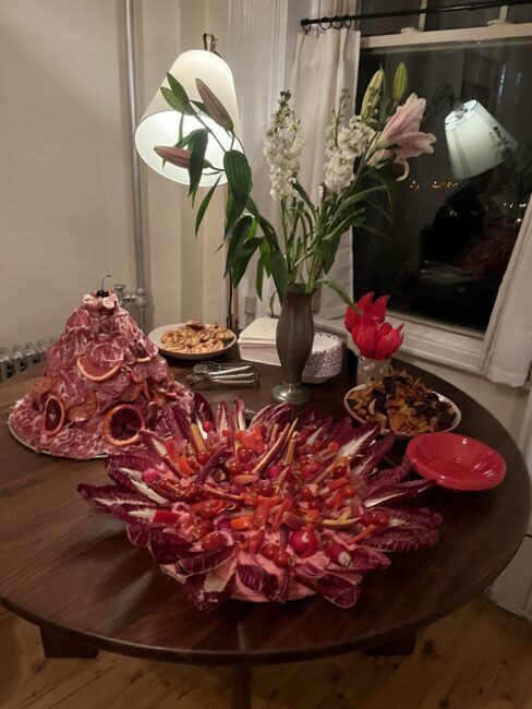 A table with a meat and radicchio platter, a meat-covered mound with citrus slices, small bowls of snacks, a vase of flowers, and a lamp, set in a dimly lit room.