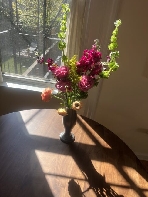 A vase of assorted flowers sits on a round wooden table in sunlight by a window, casting a shadow on the table.