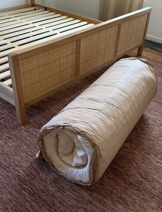 A rolled-up mattress sits on a brown rug next to a wooden bed frame with no mattress.