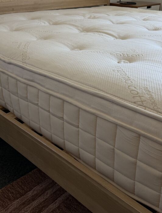 Close-up of a neatly made mattress with a quilted surface and visible stitching placed on a light wooden bed frame.