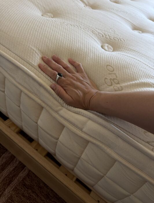 A hand pressing down on the surface of a cream-colored mattress on a wooden bed frame, showing the texture and firmness of the mattress.