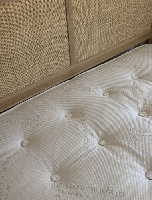 A close-up of a beige mattress labeled “organic cotton” on a wooden bed frame with a woven rattan headboard.