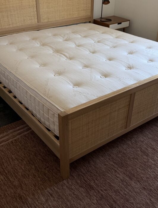 A corner view of a neatly made bed with a light wood frame, rattan panels, and a white mattress on top, placed on a reddish-brown rug in a bedroom.