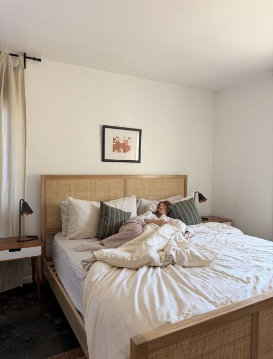 Person lying in bed under white bedding in a minimally decorated bedroom with a wooden headboard and two bedside tables with lamps.