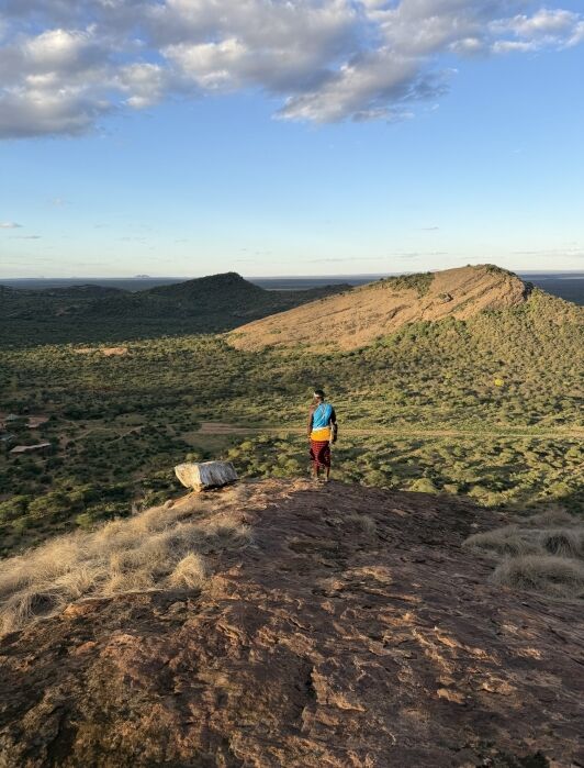 A person in colorful clothing stands on a rocky hilltop overlooking a vast, green landscape with distant hills under a partly cloudy sky.