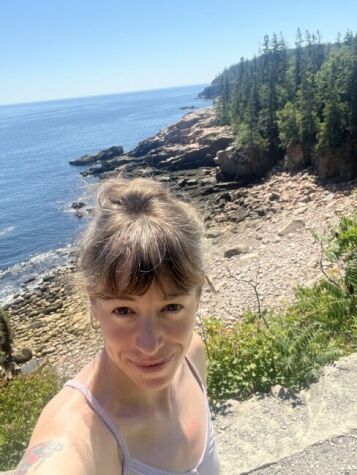 Person taking a selfie near a rocky coastline with evergreen trees, blue sky, and ocean in the background on a sunny day.