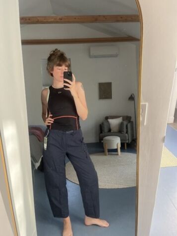Person taking a mirror selfie in a bedroom, wearing a black tank top and dark cargo pants with a tag attached. A modern chair, lamp, and rug are visible in the background.