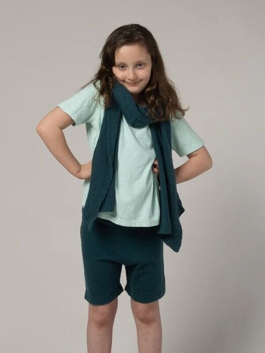 Young child with long brown hair stands with hands on hips, wearing a light blue T-shirt, dark blue scarf, and matching shorts against a plain gray background.