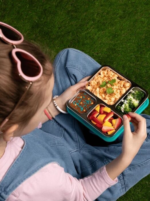 A girl holding a lunchbox with scrambled eggs, broccoli, strawberries, and orange slices sits on grass, wearing a pink shirt, denim overalls, and heart-shaped sunglasses.