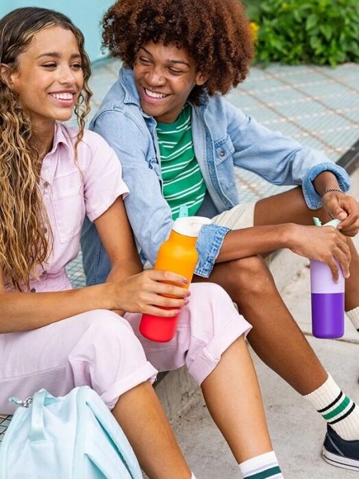 Two young people sit on outdoor steps, smiling and holding reusable water bottles; one wears pink, the other wears a striped shirt and denim jacket.