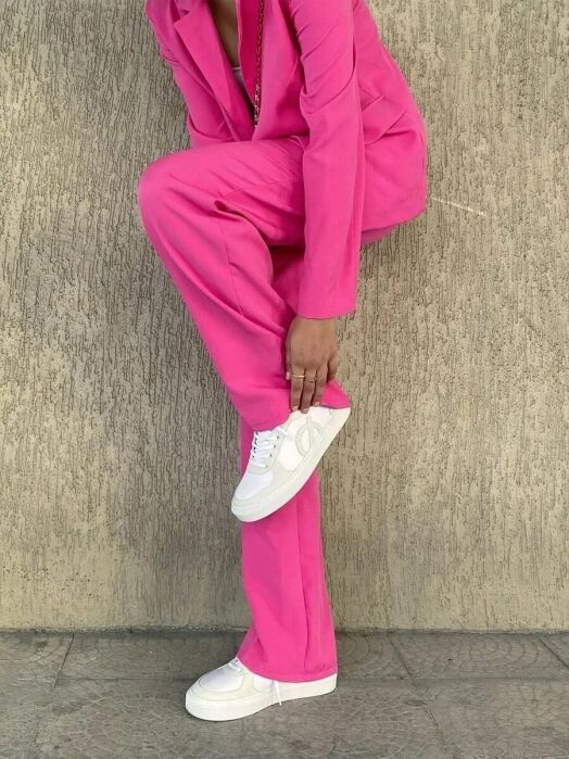 Person wearing a bright pink suit and white sneakers stands against a textured beige wall, bending forward to touch one shoe.