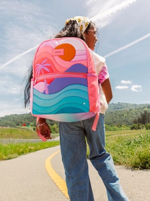 Person wearing jeans and a colorful backpack walks on a path through a green landscape under a blue sky with contrails and light clouds.