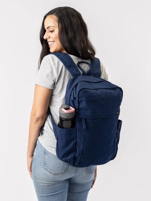 Woman wearing a gray t-shirt and jeans, carrying a navy blue backpack with a water bottle in the side pocket, standing against a plain white background.
