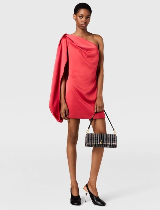 Woman wearing a one-shoulder red mini dress and black heels holds a rectangular black handbag with gold chain straps, standing against a plain light background.