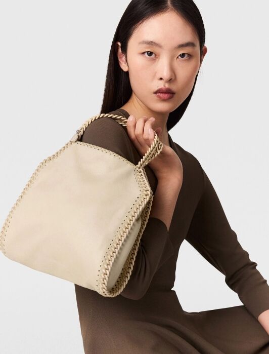 Woman with straight dark hair holds a beige handbag with braided edges and handle, wearing a long-sleeve brown dress, seated against a plain light background.