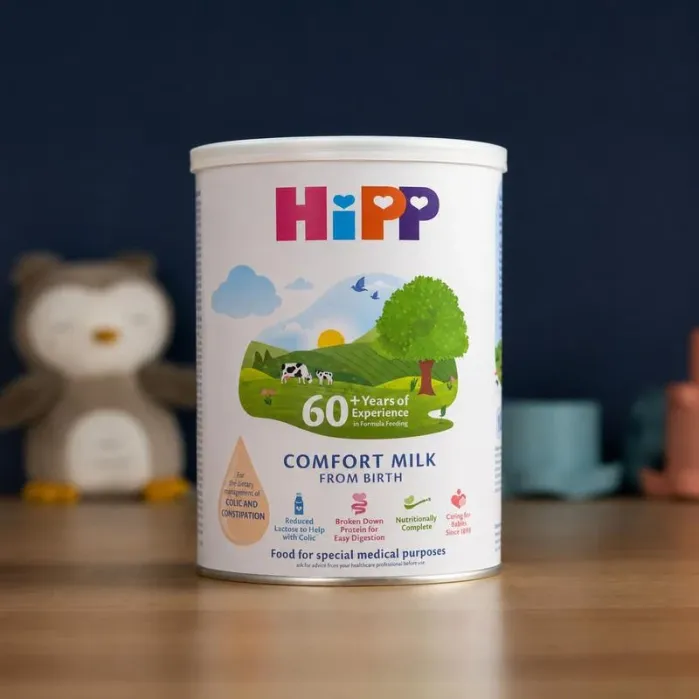 A container of HiPP Comfort Milk formula stands on a wooden surface with a blurred owl toy and other objects in the background.