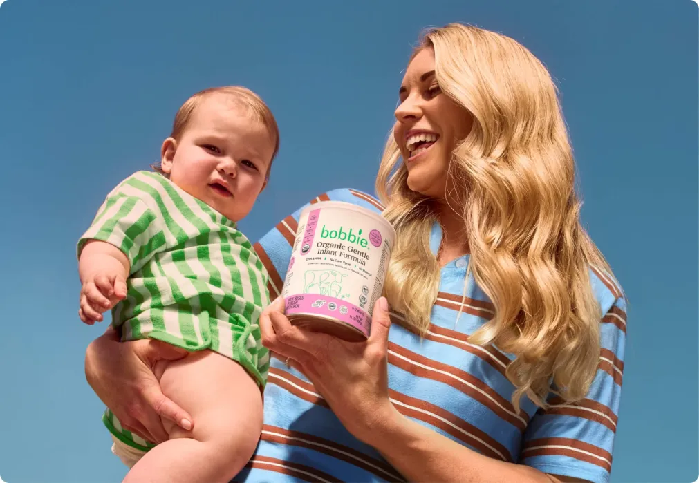 A woman holding a baby in one arm and a container of Bobbie organic infant formula in the other, both outdoors under a clear blue sky.