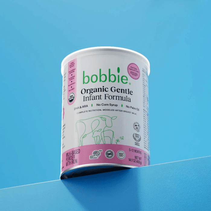 A can of Bobbie Organic Gentle Infant Formula with a white and pink label is displayed on a blue surface against a blue background.