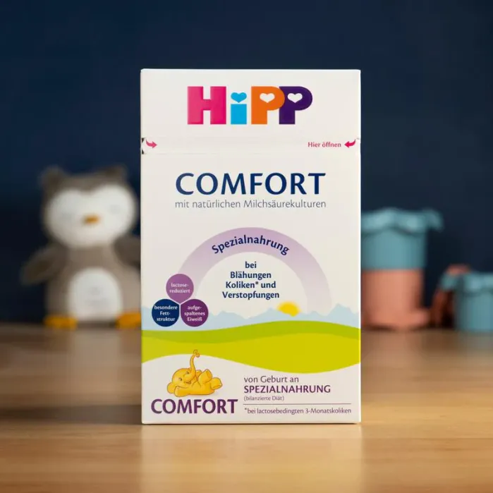 A box of HiPP Comfort infant formula stands on a wooden surface, with blurred nursery items in the background.
