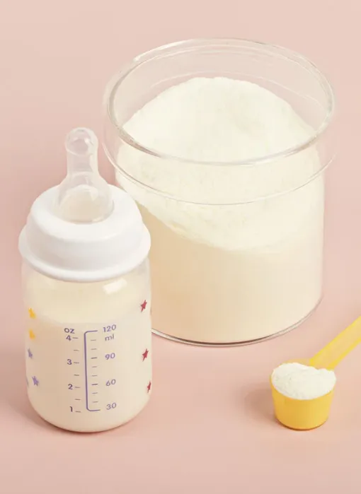 A baby bottle filled with formula, a container of powdered formula, and a yellow scoop with powdered formula on a pink background.