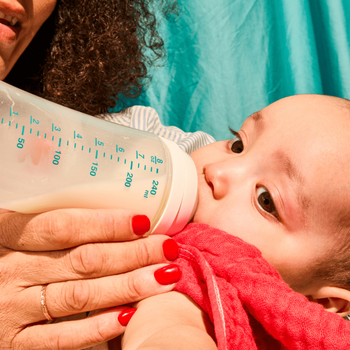 Close-up of a baby drinking milk from a bottle held by an adult with red nail polish, against a blue background.