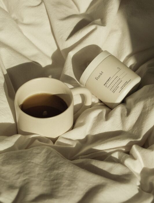 A white mug of black coffee and a white jar labeled “Pureal” rest on rumpled white bed sheets in soft sunlight.