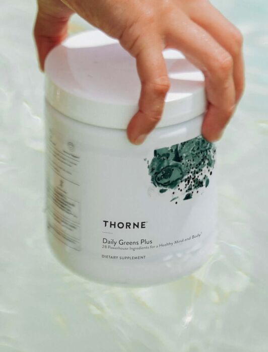 A hand holds a white Thorne Daily Greens Plus container over water. Text highlights benefits and certification of the supplement.