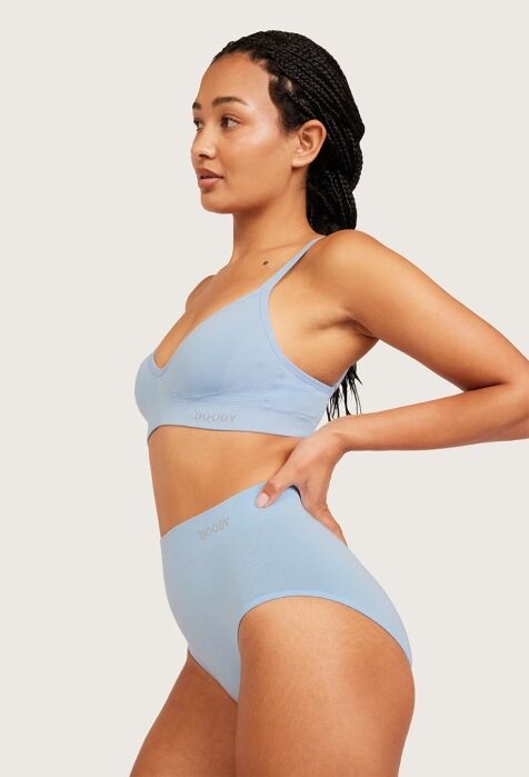 Woman wearing matching light blue bra and high-waisted underwear, standing in profile with one hand on her hip against a neutral background.