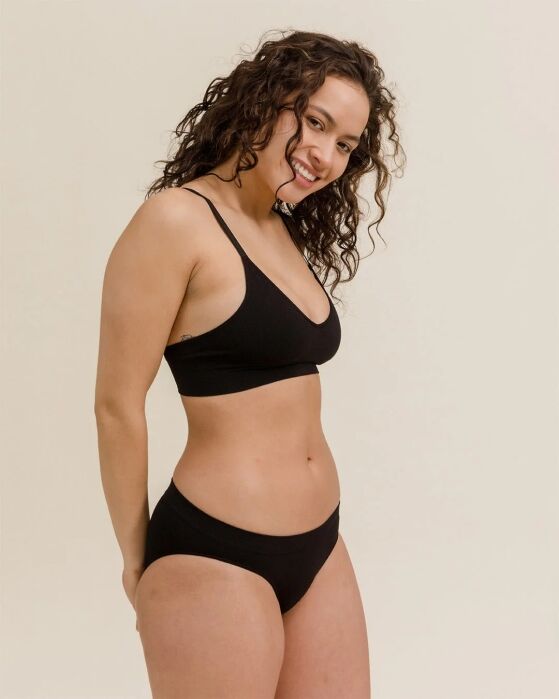 Woman with curly hair wearing a black bralette and matching underwear stands against a plain beige background, smiling at the camera.
