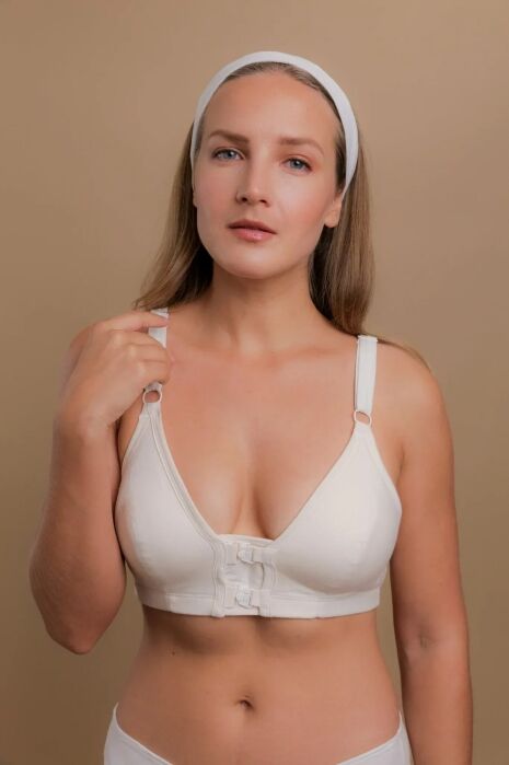 Woman wearing a white front-closure bra and matching underwear stands against a beige background, holding one strap with her right hand, looking directly at the camera.