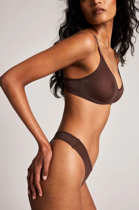 Woman wearing a brown bra and matching underwear poses with one hand on her hip against a neutral background.