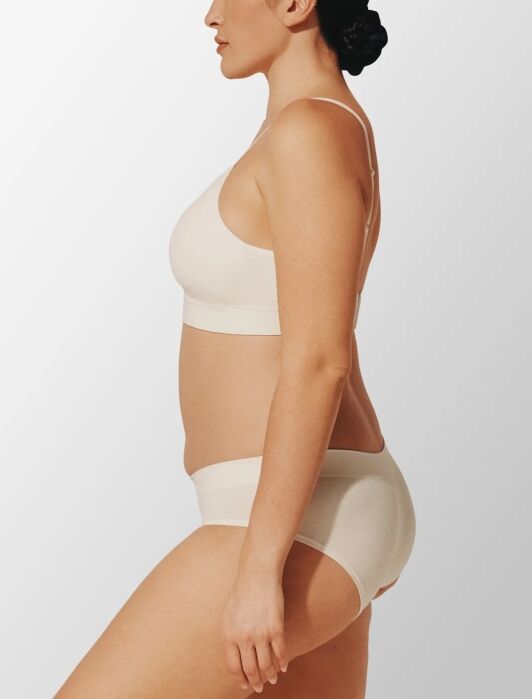 Woman in light-colored bra and underwear stands in profile against a plain background.