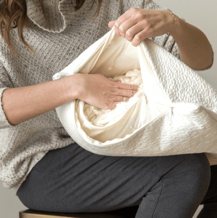 A person adjusts the filling inside a zippered pillow while sitting, wearing a gray sweater and dark pants.