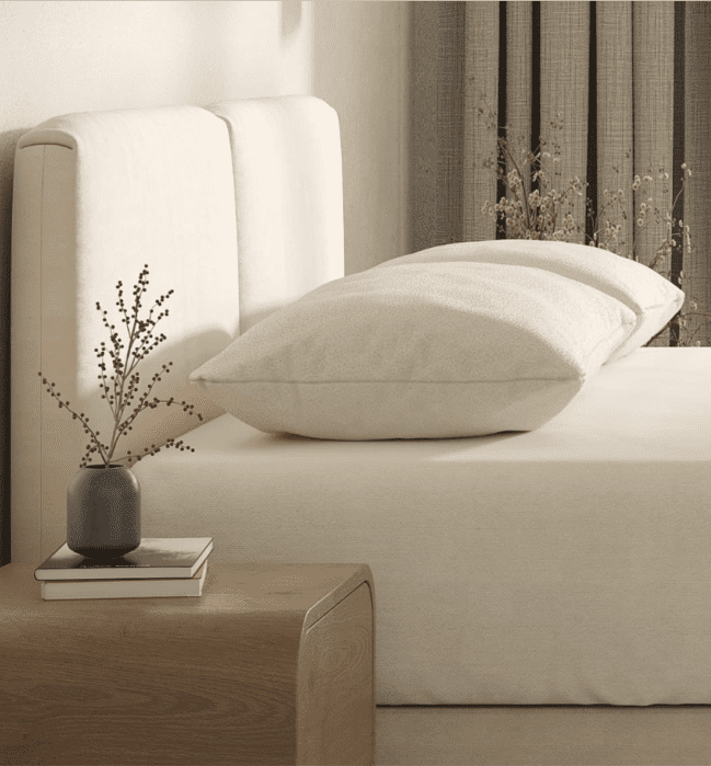 Minimalist bedroom with a beige upholstered bed, two stacked pillows, neutral bedding, a wooden nightstand, a small vase with branches, and a book.