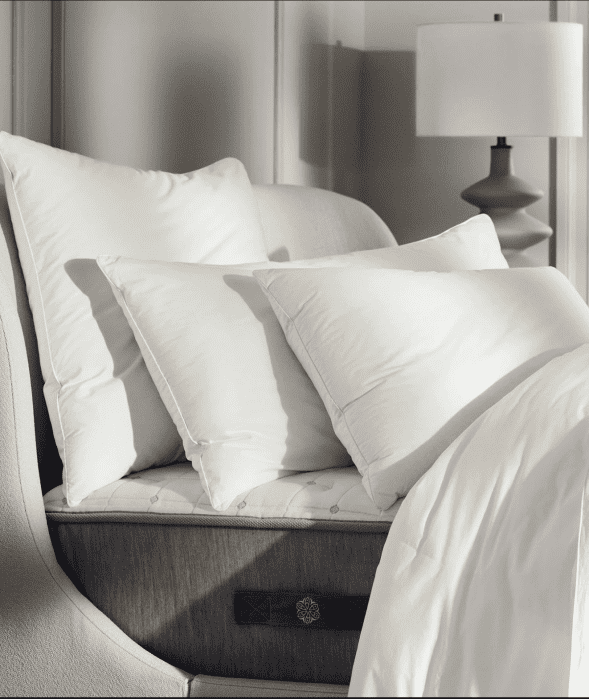 Three white pillows and a white comforter are arranged on a beige upholstered bed next to a gray table lamp.