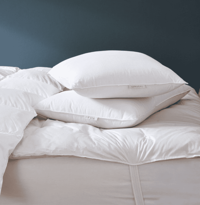 Two white pillows stacked on a white bed with a quilted comforter, set against a dark blue wall.