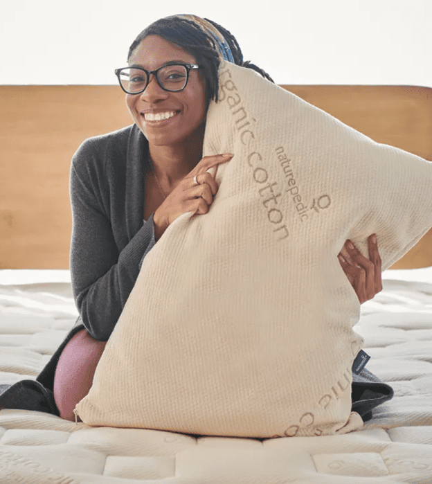 A person sits on a bed, smiling and hugging a large pillow labeled “organic cotton.”.