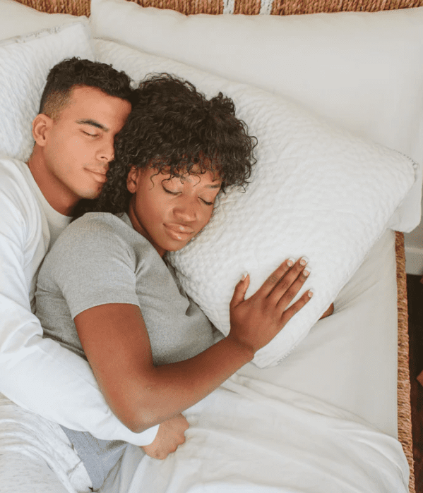 A couple lies in bed, with the man hugging the woman from behind as they both rest with their eyes closed.