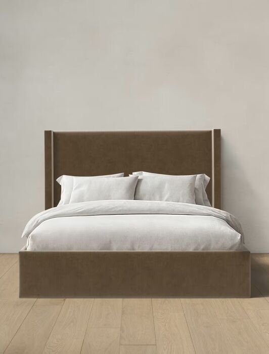 A brown upholstered bed with a high headboard, made with white bedding and pillows, sits on a light wood floor against a plain beige wall.