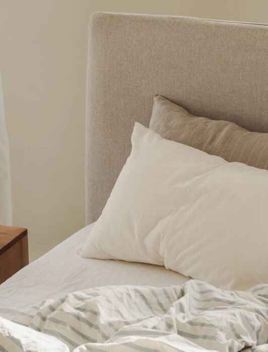 Unmade bed with white and striped bedding, two pillows, and a wooden nightstand holding books and a modern lamp in a sunlit room.
